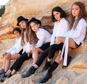 Five girls in stylish outfits sit on a sandy rock formation, showcasing a playful yet fashionable vibe in a natural outdoor setting.